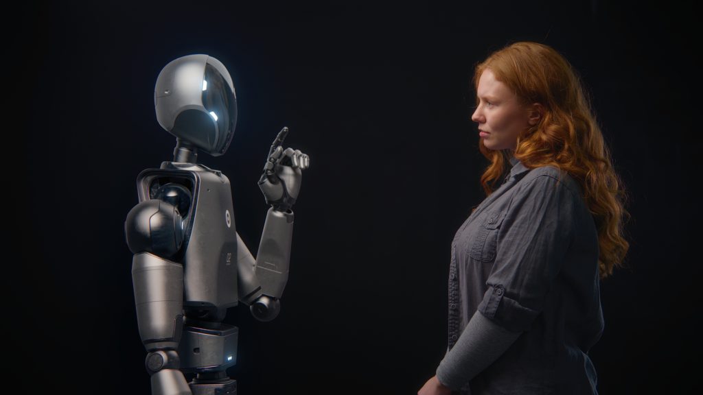 A futuristic, metallic, humanoid robot with a glowing face and a pointed finger stands facing a young woman with long, red hair. The woman, wearing a gray shirt, is looking directly at the robot with a focused expression. They are in a studio setting with a dark, uniform background.