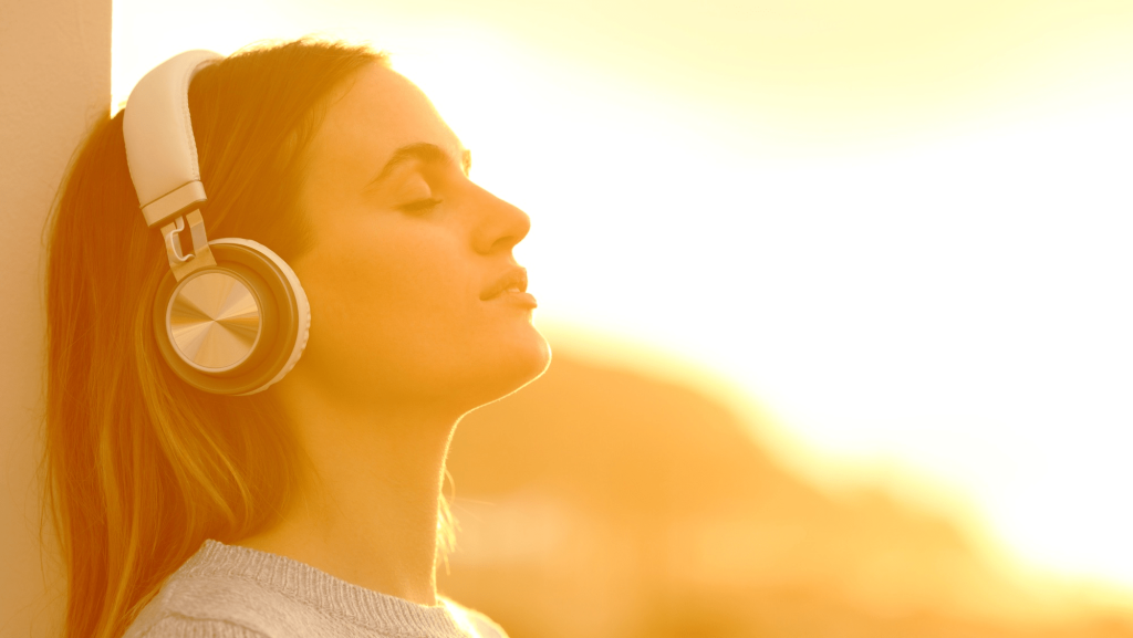 A profile shot of a woman with her eyes closed and head tilted back, leaning against a wall and wearing white over-ear headphones. She is lit by a warm, golden sunlight that creates a strong glow around her, suggesting a peaceful, contemplative moment at sunrise or sunset.